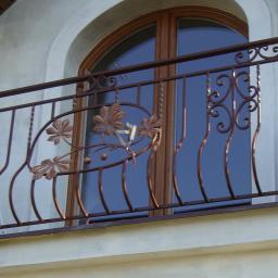 Zuh Metal-Plast - Kuta balustrada balkonowa z motywem liści kasztanowca, brązowa, na tle okna z łukiem i fragmentu dachu.